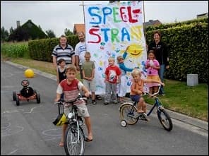 Photo d'une rue avec une banderole « Speelstraat » (rue de jeu) : des enfants à vélo et à pied, des adultes derrière eux.