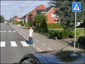 Photo d'une rue avec un passage pour piétons à l'arrière-plan : un piéton est debout au bord de la chaussée, près du passage ; le capot d'une voiture est visible au premier plan.