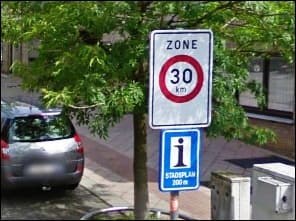 Photo d'un panneau de zone 30 au bord d'une rue, au-dessus d'un petit panneau bleu d'information ; une voiture est garée derrière.