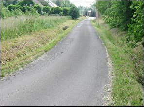 Photo d'une route de campagne étroite bordée d'herbe et de fossés, sans marquage.