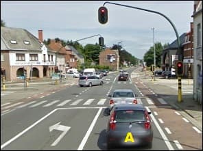 Photo d'une rue en ville : la voiture A, vue de dos, attend à des feux rouges ; à sa gauche, une bande avec une flèche vers la gauche est séparée d'elle par une ligne blanche continue.
